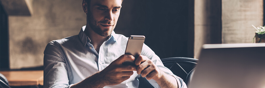 A man sitting on a desk with a laptop and looking at his mobile phone